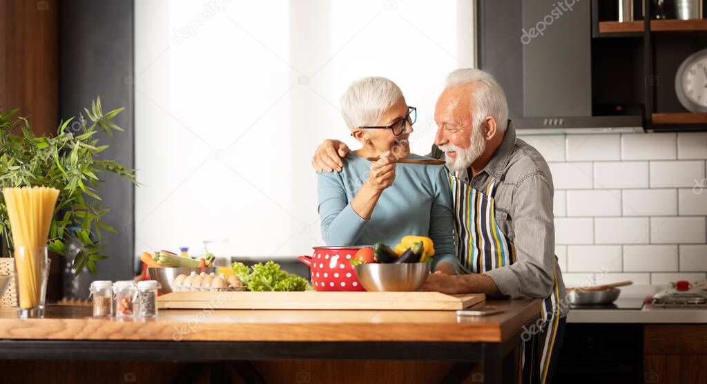 Happy senior couple cooking a meal together at home, smiling woman  feeding  his husband 