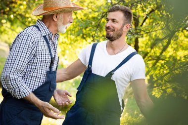 Yetişkin çiftçi oğlu meyve bahçesinde dururken büyük babasıyla konuşuyor.