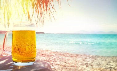 Cold glass mug of beer on beach on table under palm tree parasol clipart
