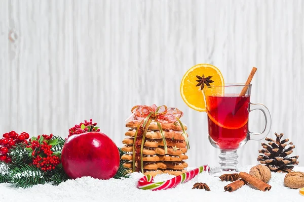 Рождественский бокал красного глинтвейна на столе с коричными палочками, ветками елки, снегом, пряниками, конусом, конфетами, новогодними украшениями на белом деревянном фоне. Свободное пространство — стоковое фото