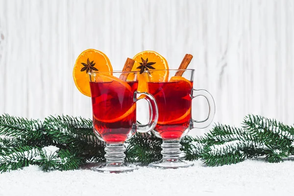 Bicchiere natalizio di vin brulè rosso su tavola con bastoncini di cannella, rami di albero di Natale, neve, pan di zenzero, cono, caramelle, decorazioni di Capodanno su sfondo bianco in legno. Spazio libero — Foto Stock