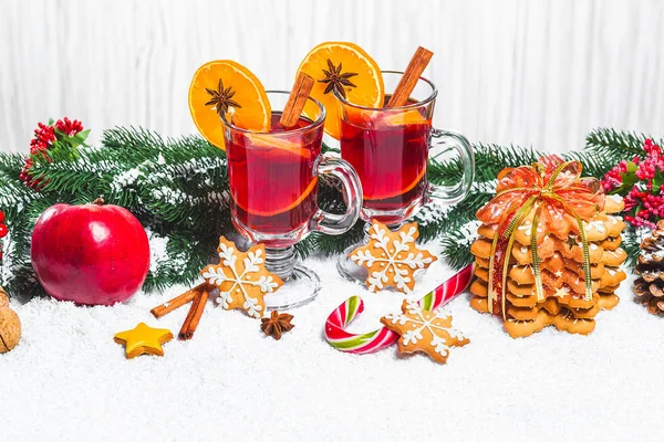 Рождественский бокал красного глинтвейна на столе с коричными палочками, ветками елки, снегом, пряниками, конусом, конфетами, новогодними украшениями на белом деревянном фоне. Свободное пространство — стоковое фото
