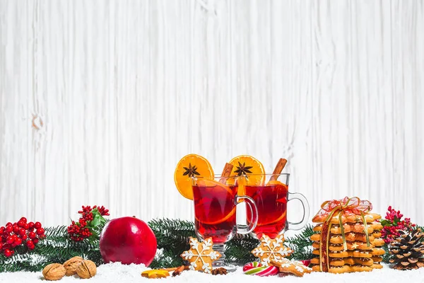 Copa de Navidad de vino tinto con especias en la mesa con palos de canela, ramas de árbol de Navidad, nieve, pan de jengibre, cono, dulces, decoraciones de Año Nuevo sobre fondo de madera blanca. Espacio libre — Foto de Stock