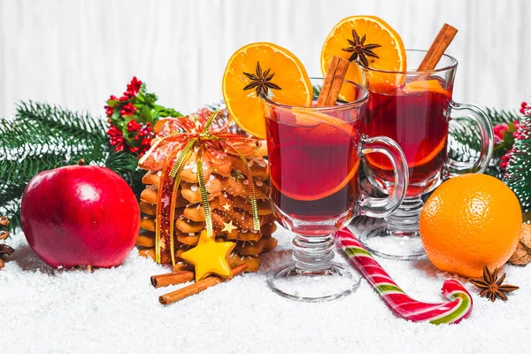 Bicchiere natalizio di vin brulè rosso su tavola con bastoncini di cannella, rami di albero di Natale, neve, pan di zenzero, cono, caramelle, decorazioni di Capodanno su sfondo bianco in legno. Spazio libero — Foto Stock