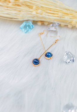 Mavi yuvarlak boncuklardan ve altın sarısı metalden yapılmış küpeler. Arka plan olarak sarı kuru çimen bitkileri. Beyaz pelüş kumaş ve şeffaf buz süslemeleri. Mavi yuvarlak boncuklar, safir taşına benzeyen taşlar, güzel dokular.. 