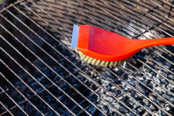 Close-up de uma escova vermelha com cerdas douradas e um raspador para limpar uma grelha de churrasco. O conceito de limpeza após o almoço ao ar livre, piquenique, churrasco, almoço — Fotografia de Stock