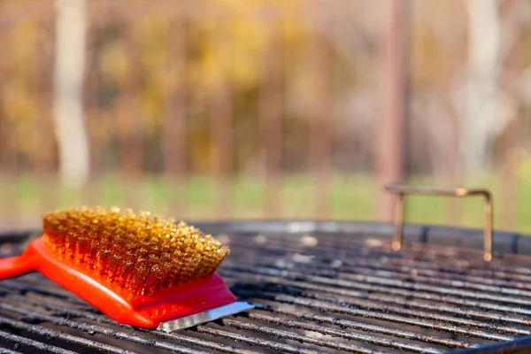 Detailní záběr červeného štětce se zlatými štětinami a škrabkou na čištění grilu. Koncept úklidu po obědě pod širým nebem, piknik, gril, oběd — Stock fotografie