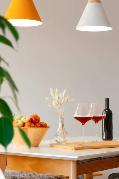 Темная бутылка и два бокала красного вина, деревянная чаша с яблоками, ваза декора на столе в современной кухне, лампы над столом, зеленые листья. По вертикали. Авторское пространство. Место для текста, интерьера — стоковое фото
