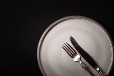 Kapalı yuvarlak, siyah arka planda gri seramik plaka, klasik cupronickel çatal, fotokopi alanı olan bıçak. Şef, restoran reklamı, sofra takımı kataloğu, üst görünüm modern çekim menüsü.