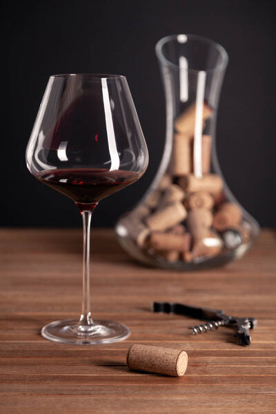 Crystal red wine glass, bottle, corkscrew, opener, sommelier knife, transparent decanter, corks on wooden table. Closeup, vertical, selective focus side view. Concept degustation winery cellar