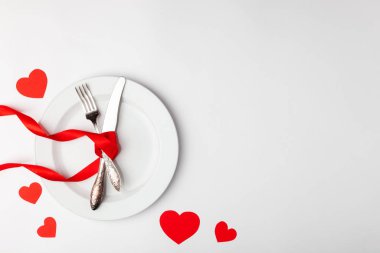 Beyaz arka planda romantik şenlik masası. Sevgililer Günü kartı şablonu. Kırmızı kurdele, tabak, çatal, çatal, bıçak. Kavram yıldönümü, doğum günü, metin yeri, telif alanı, üst görünüm