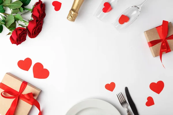 Valentines day dinner with table place setting, red tape, kraft gift box, glass for champagne, champagne bottle, roses, hearts, silverware on white background. Copyspace. Space for text. Top view