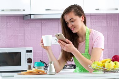 Mutlu, çekici, internette sohbet eden ve sabahın erken saatlerinde mutfakta kahvaltıda sıcak, lezzetli kahve içen bir kadın. İnternette, telefon bağımlılığı 