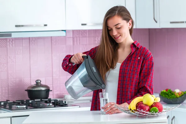 年轻女子从滤水器中倒入新鲜过滤水 倒入杯子里在厨房里喝水 家居净化水 — 图库照片