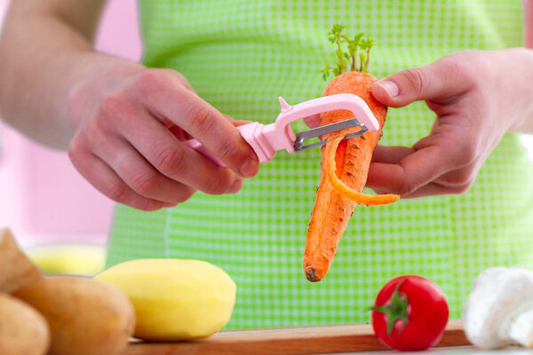 Housewife in apron peeling ripe carrot with a peeler for cooking fresh vegetable dishes and salads in kitchen at home. Proper healthy eating and clean food 