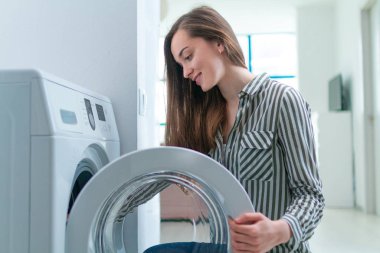 Happy housewife engaged in washing clothes and linen using washing machine at home  clipart