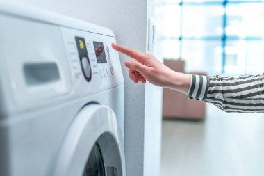 Housewife using display and button for turning on and choosing cycle program on washing machine for laundry at home. clipart