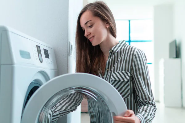 Happy housewife engaged in washing clothes and linen using washing machine at home