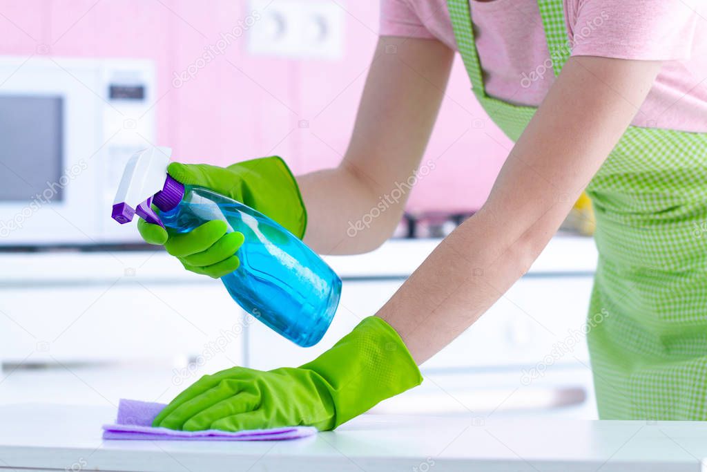 Housewife in protective rubber gloves wiping dust off the table at kitchen at home with a rag and spray. Housekeeping and household chores