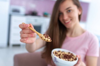 Genç, güler yüzlü, güzel esmer kadın kahvaltıda elinde mısır gevreği kaşığı ve mısır gevreği kasesi dolusu çikolata tutuyor.