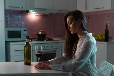 Lonely, unhappy, sad beautiful young woman in a blouse with glass of red wine is drinking alone in evening at home. Female alcoholism and alcohol addiction clipart