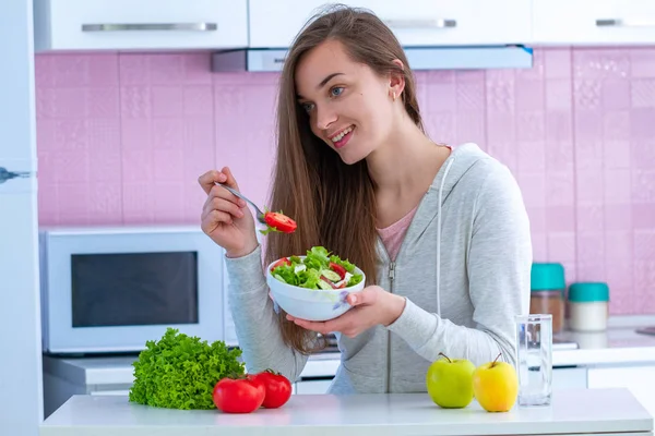 Young happy healthy woman in sportswear eating fresh vegetable salad while watching tv at home in kitchen. Balanced organic diet and clean fitness eating.