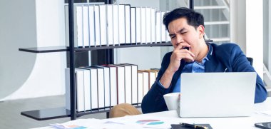 A young businessman sitting in a modern office. He has a feel sleepy because  hard work so tired weary fatigued and exhausted. On his table have a computer laptop tablet pen paper graph. clipart