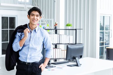 Genç Asyalı iş adamı akıllı pozlar veriyor ve yeni başlayan işletme öğrencileri ve serbest çalışanlar için modern ofis tarzında dekore edilmiş çalışma alanına gülümsüyor.