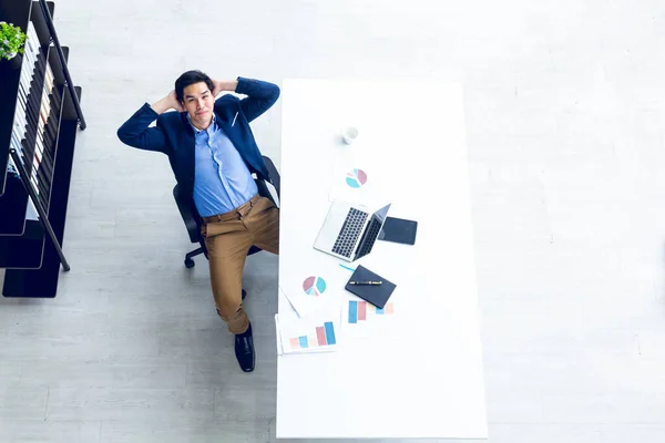 Top view of young businessman sitting in a modern office. He has a feel happy and smile about the result of business profits is positive. On his table have a computer laptop tablet pen paper graph and a coffee cup