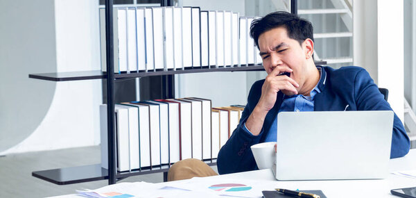 A young businessman sitting in a modern office. He has a feel sleepy because  hard work so tired weary fatigued and exhausted. On his table have a computer laptop tablet pen paper graph.