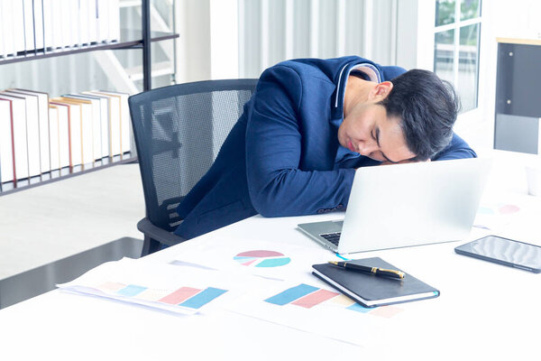 A young businessman sitting in a modern office. He has a feel sleepy because  hard work so tired weary fatigued and exhausted. On his table have a computer laptop tablet pen paper graph.