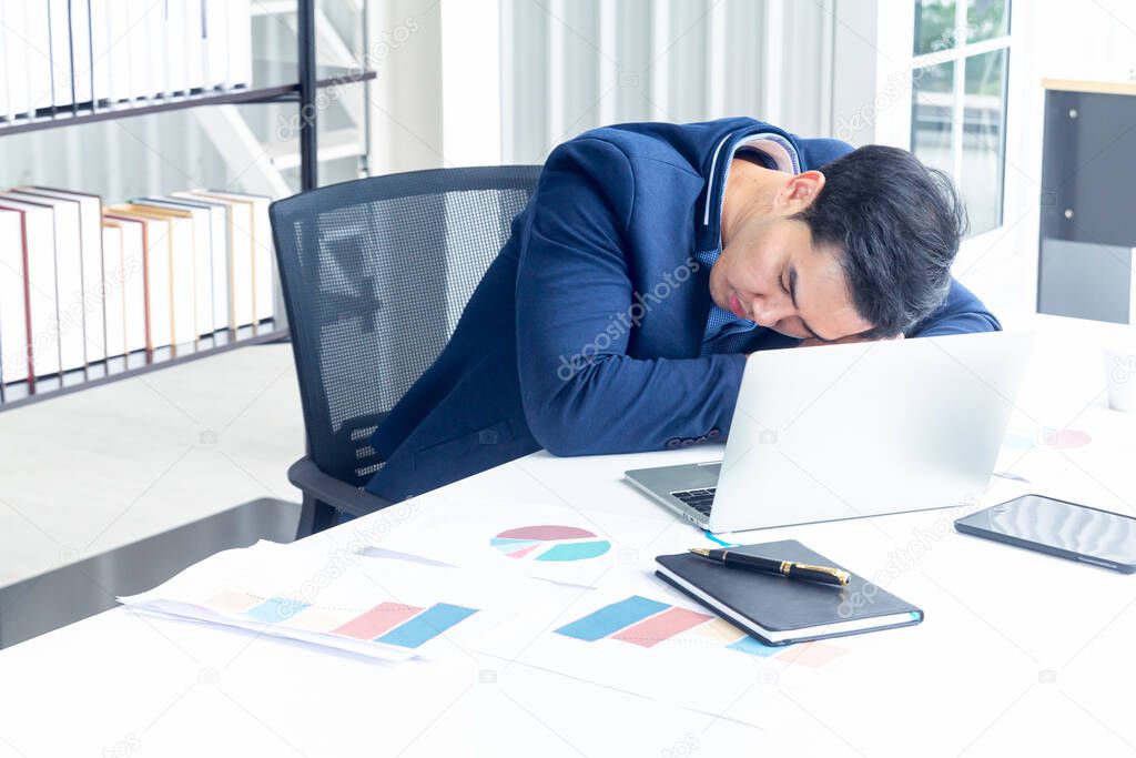 A young businessman sitting in a modern office. He has a feel sleepy because  hard work so tired weary fatigued and exhausted. On his table have a computer laptop tablet pen paper graph.