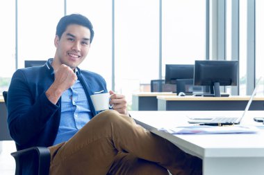 Takım elbiseli, uzun kollu gömlekli ve şık pozlar veren genç bir işadamının modern bir ofiste mutlu bir gülümsemesi vardır. Masasında bir bilgisayar tableti kağıt kalem grafiği ve elinde bir kahve fincanı var..