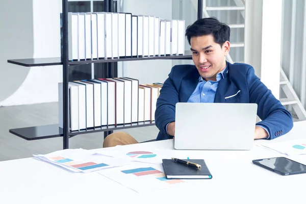 Top view of young businessman sitting in a modern office. He has a feel happy and smile about the result of business profits is positive. On his table have a computer laptop tablet pen paper graph and a coffee cup