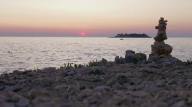 Pembe deniz günbatımına ve karanlık adaya karşı taştan kumsalda yelkenliler ve çimler. Alacakaranlıkta sudaki yelkenli silueti, akşam renkli gökyüzü. Yol işareti ve sürat teknesi olarak çakıl taşı kulesi. Adriyatik 'te romantik aktif tatil