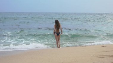 İnce bir kız okyanus boyunca yürür ve ayaklarını suda kurutur. Kulaklıkla akıllı bir telefondan müzik dinler.