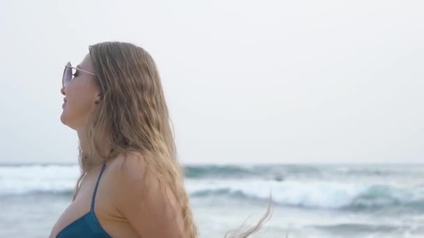 Όμορφη γυναίκα με μακριά μαλλιά σε γυαλιά, βόλτες κατά μήκος του ωκεανού, χαμόγελα, χοροί, ακούει μουσική, αργή κίνηση — Αρχείο Βίντεο