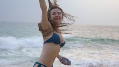 Uzun saçlı güzel bir kız, kumsalda yürüyor ve akıllı telefondan müzik dinliyor ve dans ediyor.