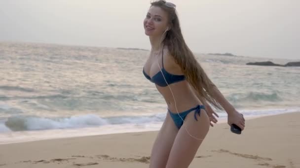 Egy karcsú lány hosszú hajjal sétál az óceán mentén, táncol, zenét hallgat. — Stock videók