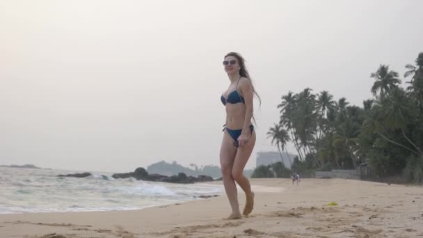 Mooi meisje met lang haar in bril, wandelingen langs de oceaan, glimlacht, danst, luistert naar muziek, slow motion — Stockvideo