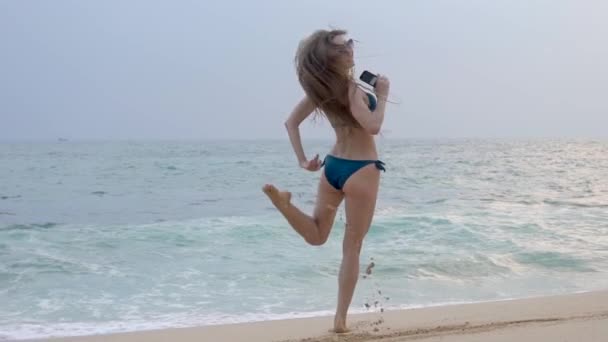 Schönes Mädchen mit langen Haaren und Brille, geht am Meer entlang, lächelt, tanzt, hört Musik, Zeitlupe — Stockvideo