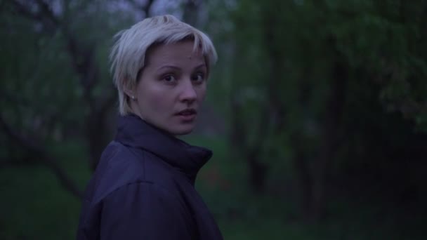Mädchen, blond, mit Kurzhaarschnitt, in schwarzer Jacke, spaziert spätabends im Garten — Stockvideo