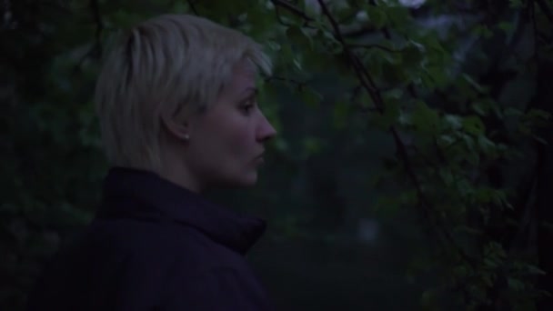 검은 재킷을 입고 짧은 머리를 한 금발의 여자가 늦은 저녁에 정원에서 산책 — 비디오