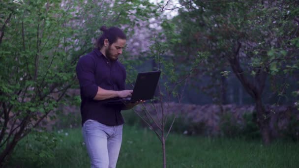 Uzun saçlı, mavi gömlekli, kot pantolonlu, dizüstü bilgisayarı olan bir adam bahçede. — Stok video
