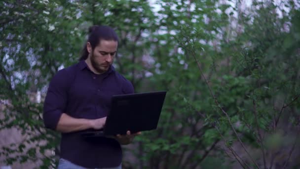 Ein Typ mit Laptop, langen Haaren geht durch den Garten und tippt Text — Stockvideo