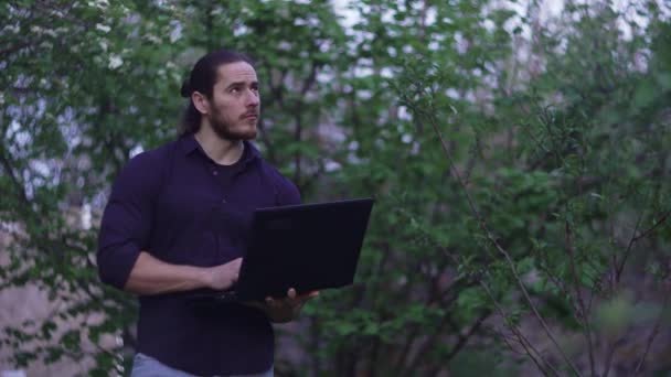 Een man met lang haar in een blauw shirt en spijkerbroek, wandelt in de tuin — Stockvideo