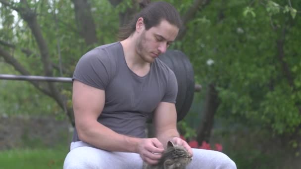 Hombre atleta se sienta en un simulador de deportes en su patio y acaricia a un gato — Vídeos de Stock