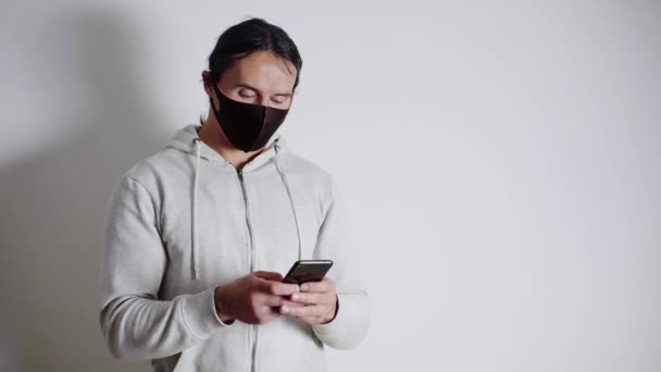 Un giovane uomo con una maschera medica nera,, con un maglione bianco, scrive SMS al telefono, guarda lo smartphone — Video Stock