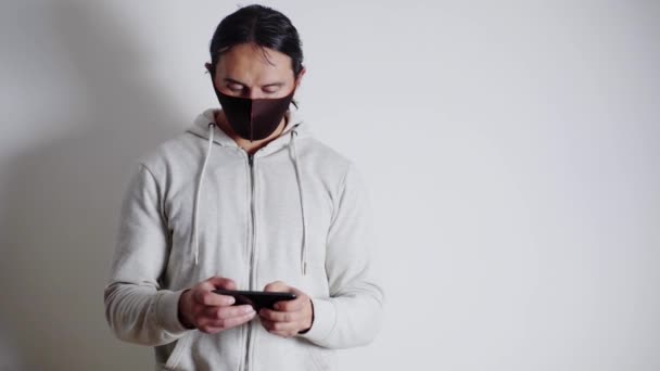 스스로 고립되고 격리되고 의료용 마스크를 쓴 젊은이가 스마트폰으로 게임을 하고 있습니다. — 비디오