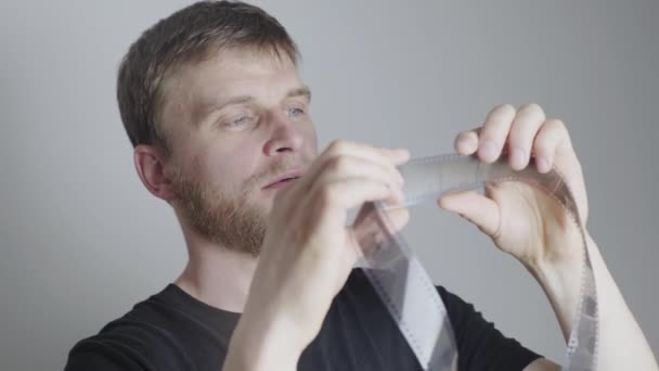 Um homem loiro com barba, um fotógrafo, examina um filme, sobre um fundo branco — Vídeo de Stock